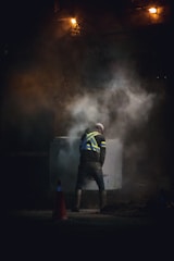A construction worker in a high-visibility vest stands amidst rising smoke or dust, illuminated by a dim light. The scene appears to be taken at night or in a low-light environment, adding to the dramatic effect. Traffic cones are visible on the ground.