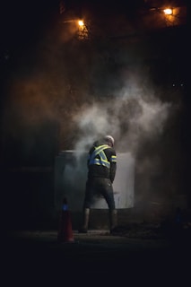 A construction worker in a high-visibility vest stands amidst rising smoke or dust, illuminated by a dim light. The scene appears to be taken at night or in a low-light environment, adding to the dramatic effect. Traffic cones are visible on the ground.