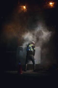 A construction worker in a high-visibility vest stands amidst rising smoke or dust, illuminated by a dim light. The scene appears to be taken at night or in a low-light environment, adding to the dramatic effect. Traffic cones are visible on the ground.