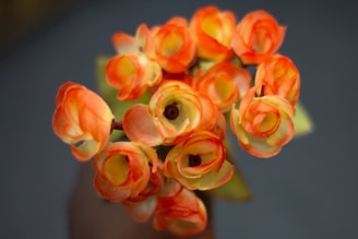 A close-up view of several small artificial flowers with vibrant orange petals and green centers. The petals appear to be crafted from fabric, capturing the soft and delicate texture typical of imitation florals. The background is softly out of focus, emphasizing the vivid colors and details of the floral arrangement.