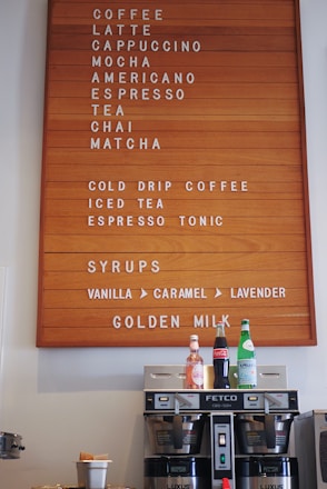 A wooden menu board displays various beverages including coffee, latte, cappuccino, mocha, americano, espresso, tea, chai, matcha, and more. Below the menu are syrups like vanilla, caramel, and lavender. Beneath the board are a coffee machine and three bottles of different drinks including sparkling water and cola.