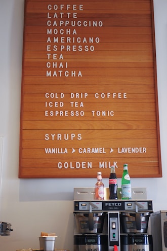 A wooden menu board displays various beverages including coffee, latte, cappuccino, mocha, americano, espresso, tea, chai, matcha, and more. Below the menu are syrups like vanilla, caramel, and lavender. Beneath the board are a coffee machine and three bottles of different drinks including sparkling water and cola.