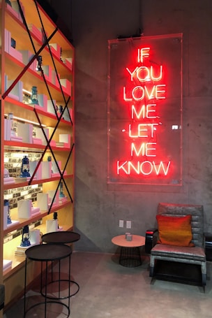 A cozy corner with a concrete wall and a neon sign reading 'IF YOU LOVE ME LET ME KNOW' in red letters. A leather chair with an orange and brown cushion is positioned next to a small round table. Shelves filled with books and blue lanterns occupy the left side of the image, alongside two nested round tables.