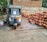 A weathered autorickshaw is parked in front of a stack of clay pots. The vehicle appears old, with signs of rust and wear. The background includes a stone wall and a brick structure. There is a container with paint or some liquid next to the autorickshaw.