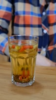 A glass filled with a yellowish beverage containing floating herbs and small red berries or fruits, placed on a wooden surface. The background shows a person wearing a multicolored, checkered shirt in shades of orange, blue, and beige.
