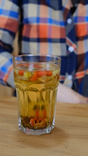 A glass filled with a yellowish beverage containing floating herbs and small red berries or fruits, placed on a wooden surface. The background shows a person wearing a multicolored, checkered shirt in shades of orange, blue, and beige.