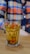 A glass filled with a yellowish beverage containing floating herbs and small red berries or fruits, placed on a wooden surface. The background shows a person wearing a multicolored, checkered shirt in shades of orange, blue, and beige.