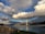 A large bridge spans across a body of water under a dramatic sky filled with diverse and vibrant cloud formations. The bridge features modern architectural elements, and the water below reflects the clouds and the structure. In the foreground, there is a dirt path with a few scattered barrels, which leads towards the water's edge. On the left side, trees and a hint of hills can be seen in the distance.