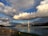 A large bridge spans across a body of water under a dramatic sky filled with diverse and vibrant cloud formations. The bridge features modern architectural elements, and the water below reflects the clouds and the structure. In the foreground, there is a dirt path with a few scattered barrels, which leads towards the water's edge. On the left side, trees and a hint of hills can be seen in the distance.