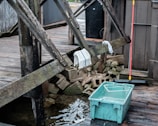 A wooden deck with worn beams, a couple of towels draped over them, and a stack of concrete blocks beneath. A blue plastic container filled with water sits on the wet wooden surface near the deck. A red-handled broom is leaning against the structure, adding a hint of activity or recent use.
