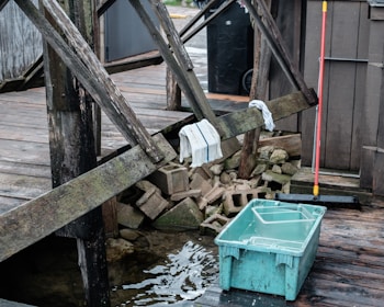 A wooden deck with worn beams, a couple of towels draped over them, and a stack of concrete blocks beneath. A blue plastic container filled with water sits on the wet wooden surface near the deck. A red-handled broom is leaning against the structure, adding a hint of activity or recent use.