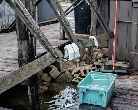 A wooden deck with worn beams, a couple of towels draped over them, and a stack of concrete blocks beneath. A blue plastic container filled with water sits on the wet wooden surface near the deck. A red-handled broom is leaning against the structure, adding a hint of activity or recent use.
