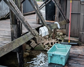 A wooden deck with worn beams, a couple of towels draped over them, and a stack of concrete blocks beneath. A blue plastic container filled with water sits on the wet wooden surface near the deck. A red-handled broom is leaning against the structure, adding a hint of activity or recent use.