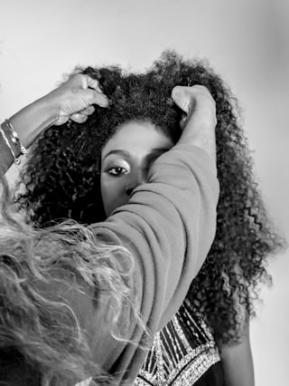 A person with curly hair is having their hair adjusted by another person. The setting appears intimate, possibly a hair salon or a styling session. The image is in black and white, highlighting textures and contrasts.