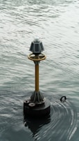 A navigational buoy floats on a body of water, featuring solar panels and a light on top. The surrounding water displays subtle ripples and a muted, overcast reflection.
