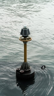 A navigational buoy floats on a body of water, featuring solar panels and a light on top. The surrounding water displays subtle ripples and a muted, overcast reflection.