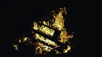 A pile of wooden pallets engulfed in bright, swirling flames against a dark background. The fire creates an intense and dramatic contrast with the darkness, with flames prominently showcasing shades of yellow and orange.