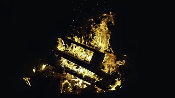 A pile of wooden pallets engulfed in bright, swirling flames against a dark background. The fire creates an intense and dramatic contrast with the darkness, with flames prominently showcasing shades of yellow and orange.