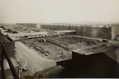 A large industrial complex with long warehouse buildings and rows of material piles, likely coal or gravel. The structures are arranged in a grid pattern with multiple access roads and pathways. A few vehicles are visible, including trucks, near the materials, indicating activity in the area. The landscape is flat with a distant horizon, and the photograph appears to be an aerial or elevated view.