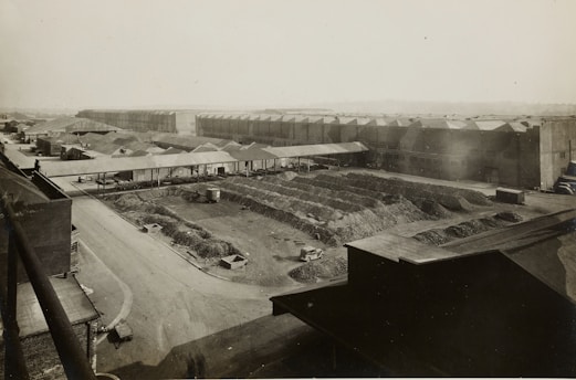 A large industrial complex with long warehouse buildings and rows of material piles, likely coal or gravel. The structures are arranged in a grid pattern with multiple access roads and pathways. A few vehicles are visible, including trucks, near the materials, indicating activity in the area. The landscape is flat with a distant horizon, and the photograph appears to be an aerial or elevated view.