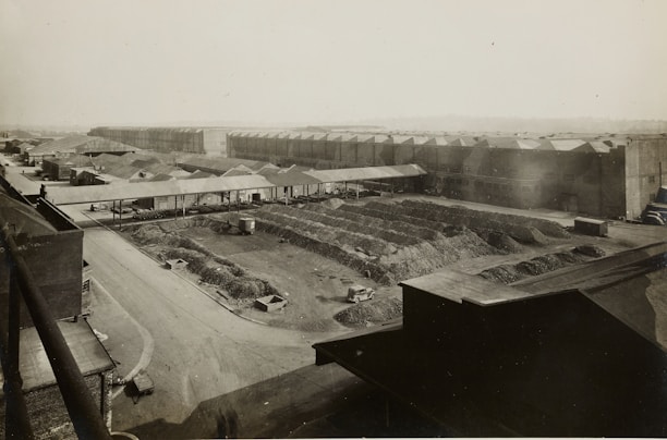 A large industrial complex with long warehouse buildings and rows of material piles, likely coal or gravel. The structures are arranged in a grid pattern with multiple access roads and pathways. A few vehicles are visible, including trucks, near the materials, indicating activity in the area. The landscape is flat with a distant horizon, and the photograph appears to be an aerial or elevated view.