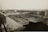 A large industrial complex with long warehouse buildings and rows of material piles, likely coal or gravel. The structures are arranged in a grid pattern with multiple access roads and pathways. A few vehicles are visible, including trucks, near the materials, indicating activity in the area. The landscape is flat with a distant horizon, and the photograph appears to be an aerial or elevated view.
