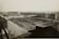 A large industrial complex with long warehouse buildings and rows of material piles, likely coal or gravel. The structures are arranged in a grid pattern with multiple access roads and pathways. A few vehicles are visible, including trucks, near the materials, indicating activity in the area. The landscape is flat with a distant horizon, and the photograph appears to be an aerial or elevated view.