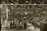 A large industrial factory setting is filled with numerous workers busy at their stations. The space is organized with rows of workbenches and various industrial machinery. Overhead lighting illuminates the expansive area, while large signs hang from the ceiling indicating different sections such as 'Bay D.2 D.3'. The scene conveys a bustling atmosphere typical of mid-20th century manufacturing environments.