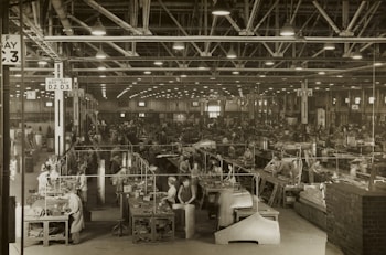 A large industrial factory setting is filled with numerous workers busy at their stations. The space is organized with rows of workbenches and various industrial machinery. Overhead lighting illuminates the expansive area, while large signs hang from the ceiling indicating different sections such as 'Bay D.2 D.3'. The scene conveys a bustling atmosphere typical of mid-20th century manufacturing environments.