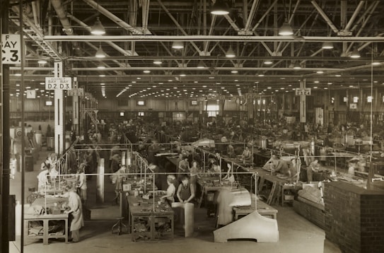 A large industrial factory setting is filled with numerous workers busy at their stations. The space is organized with rows of workbenches and various industrial machinery. Overhead lighting illuminates the expansive area, while large signs hang from the ceiling indicating different sections such as 'Bay D.2 D.3'. The scene conveys a bustling atmosphere typical of mid-20th century manufacturing environments.