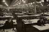 A large industrial workspace filled with numerous workers seated at drafting tables spread across the room. The environment appears busy and orderly, with workers focused on papers and tools. The architecture is reminiscent of a factory or warehouse with high ceilings and rows of symmetrical windows and lights.