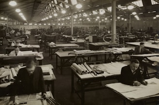 A large industrial workspace filled with numerous workers seated at drafting tables spread across the room. The environment appears busy and orderly, with workers focused on papers and tools. The architecture is reminiscent of a factory or warehouse with high ceilings and rows of symmetrical windows and lights.