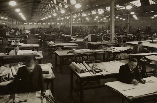 A large industrial workspace filled with numerous workers seated at drafting tables spread across the room. The environment appears busy and orderly, with workers focused on papers and tools. The architecture is reminiscent of a factory or warehouse with high ceilings and rows of symmetrical windows and lights.
