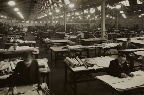 A large industrial workspace filled with numerous workers seated at drafting tables spread across the room. The environment appears busy and orderly, with workers focused on papers and tools. The architecture is reminiscent of a factory or warehouse with high ceilings and rows of symmetrical windows and lights.