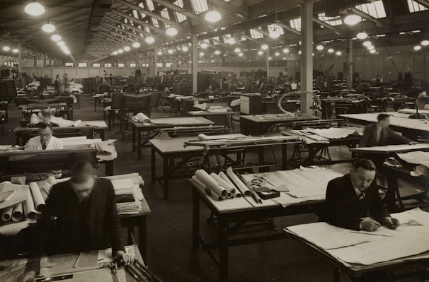 A large industrial workspace filled with numerous workers seated at drafting tables spread across the room. The environment appears busy and orderly, with workers focused on papers and tools. The architecture is reminiscent of a factory or warehouse with high ceilings and rows of symmetrical windows and lights.