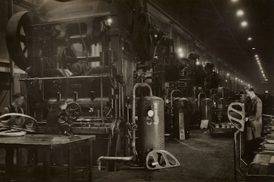 A large industrial factory setting with heavy machinery and equipment. Several workers are engaged with the machines, appearing to be operating or maintaining them. The scene is dimly lit, with numerous overhead lights leading into the distance. The machinery is robust and complex, indicating a heavy manufacturing process.