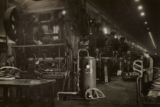 A large industrial factory setting with heavy machinery and equipment. Several workers are engaged with the machines, appearing to be operating or maintaining them. The scene is dimly lit, with numerous overhead lights leading into the distance. The machinery is robust and complex, indicating a heavy manufacturing process.