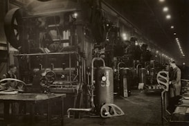 A large industrial factory setting with heavy machinery and equipment. Several workers are engaged with the machines, appearing to be operating or maintaining them. The scene is dimly lit, with numerous overhead lights leading into the distance. The machinery is robust and complex, indicating a heavy manufacturing process.