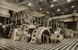 A large industrial space with massive machinery, including engines and generators. Two workers are operating and maintaining the equipment on a checkered floor under a high ceiling with steel beams.