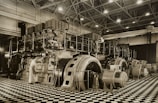 A large industrial space with massive machinery, including engines and generators. Two workers are operating and maintaining the equipment on a checkered floor under a high ceiling with steel beams.