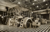 A large industrial space with massive machinery, including engines and generators. Two workers are operating and maintaining the equipment on a checkered floor under a high ceiling with steel beams.
