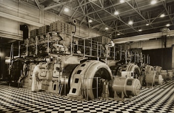 A large industrial space with massive machinery, including engines and generators. Two workers are operating and maintaining the equipment on a checkered floor under a high ceiling with steel beams.