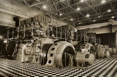 A large industrial space with massive machinery, including engines and generators. Two workers are operating and maintaining the equipment on a checkered floor under a high ceiling with steel beams.