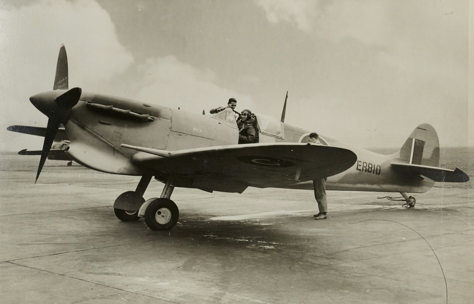 A vintage military aircraft is stationed on an airfield. Two people are interacting with the plane; one is sitting inside the cockpit while the other is standing on the ground, close to the aircraft. The plane is a single-propeller type, with distinctive markings and numbers visible on the fuselage.