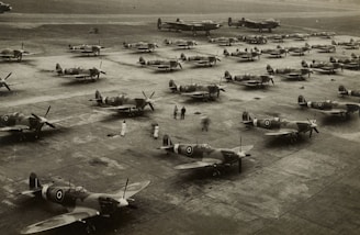 A large number of military aircraft are lined up on a tarmac in an organized manner, suggesting preparation or storage. People are seen walking around the planes, possibly performing inspections or maintenance. The image, in black and white, gives a historical feel.