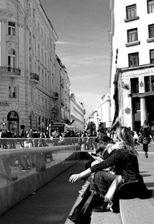 A bustling city street with crowds of people walking and socializing. Two women are sitting on steps, engaged with their phones, while others pass by. Architectural details of the historical buildings frame the scene, capturing urban life in motion.