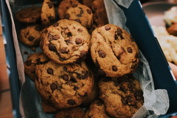 A box of freshly baked chocolate chip cookies is arranged neatly, with visible chocolate chunks scattered throughout the golden brown surface of each cookie.