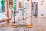 A sculpture made of wire mesh sits on a wooden swing suspended by ropes. The figure appears to be transparent and is intricately crafted to resemble a human form with an ethereal appearance. The room in the background features a polished concrete floor, large windows showing greenery outside, and various artworks displayed on the walls, creating an open and artistic atmosphere.