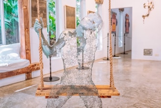 A sculpture made of wire mesh sits on a wooden swing suspended by ropes. The figure appears to be transparent and is intricately crafted to resemble a human form with an ethereal appearance. The room in the background features a polished concrete floor, large windows showing greenery outside, and various artworks displayed on the walls, creating an open and artistic atmosphere.