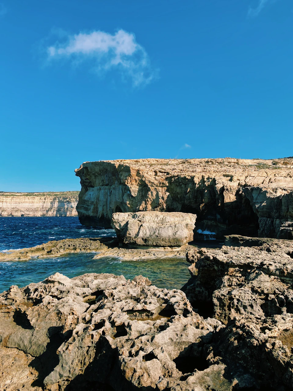 The image depicts a rocky cliff overlooking the ocean, with a clear blue sky above and white clouds scattered across it. The cliff is composed of large rocks and boulders, creating a rugged and natural landscape. In the foreground, there are small waves crashing against the shoreline, adding a dynamic element to the scene.